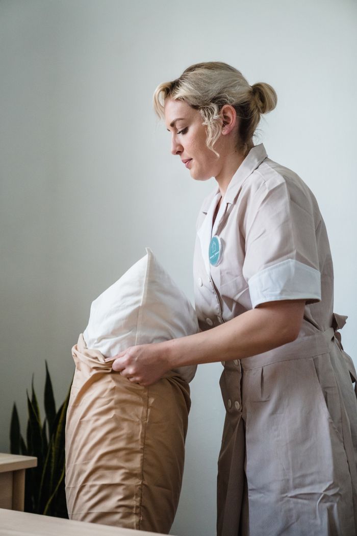 A hotel maid in uniform is arranging pillows in a neatly styled guest room.